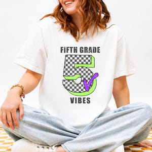 May include: A white short-sleeved t-shirt featuring a graphic design with the text FIFTH GRADE VIBES in dark gray. The center displays a large number 5 with a black and white checkered pattern, a bright green outline, and a purple checkmark.