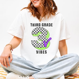 May include: A white short-sleeved t-shirt featuring a graphic with the text THIRD GRADE VIBES in black. The center displays a large number 3 with a black and white checkered pattern, a lime green outline, and a purple checkmark accent.