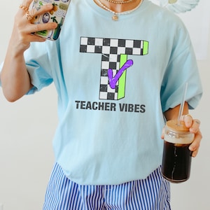 May include: Light blue t-shirt with a large, checkered "T" design outlined in green and a purple checkmark. The text "TEACHER VIBES" is printed below the design in black. The shirt is made of a soft material.