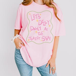 May include: A light pink short-sleeved t-shirt featuring a wavy-edged sage green border enclosing the pink text "LET'S DILLY DALLY AT THE THRIFT STORE" accented with small yellow four-pointed star shapes scattered throughout the central graphic design.