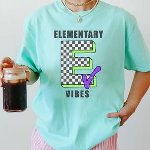 May include: A light mint green short-sleeve t-shirt featuring the text "ELEMENTARY VIBES" in dark gray. The center graphic displays a large letter "E" with a black and white checkerboard pattern, a bright green outline, and a purple checkmark.