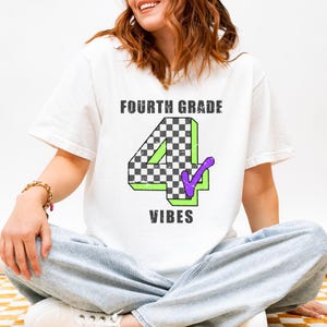 May include: A white short-sleeved t-shirt featuring the text "FOURTH GRADE" in black, a large number "4" with a black and white checkered pattern and a green outline, a purple checkmark, and the word "VIBES" in black lettering below the number.