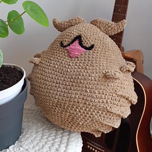 May include: A handmade, tan crochet plushie with a pink nose and black mustache. The plushie has two small horns and is resting on a white surface. A brown acoustic guitar is in the background.