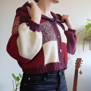 Op de afbeelding: Een kort vest met capuchon in een color-block patroon van donkerbordeaux, felrood en crème. Het gebreide kledingstuk heeft lange mouwen met geribbelde boorden, een geribbelde zoom en een knoopsluiting aan de voorzijde met vier donkerbruine knopen.