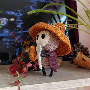 Può includere: Una figura amigurumi all'uncinetto con un grande cappello arancione a tesa larga, una maschera beige con fori neri per gli occhi, un corpo marrone e un mantello viola scuro. La figura tiene in mano un piccolo oggetto chiaro a forma di cucchiaio.
