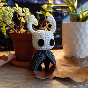 May include: A handmade crochet figure of a character with a white head, black eyes, and a grey cape. The figure is placed on a wooden surface with plants in the background. The figure is approximately 10 cm tall.