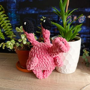 May include: A handmade pink crochet flamingo with black accents and a textured appearance. The flamingo is placed on a wooden surface, next to potted plants with green foliage and a white pot.