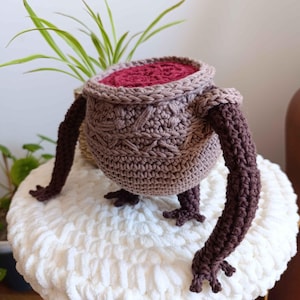 May include: A handcrafted crochet cauldron in shades of brown and mauve, featuring long, dark brown arms and feet. The cauldron rests on a white, fluffy surface, with a plant in the background. The interior of the cauldron is a deep burgundy colour.