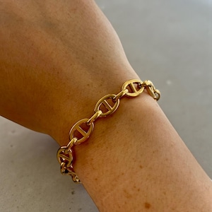 Op de afbeelding: Een goudkleurige schakelarmband. De armband heeft een reeks ovaalvormige schakels die in het midden door een staafje zijn verbonden. De armband wordt om een pols gedragen en lijkt van metaal te zijn.