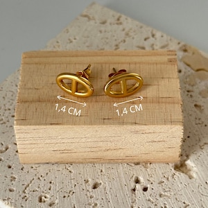 May include: A pair of gold-colored oval stud earrings with a vertical bar design. Each earring measures 0.55 inches in length. The earrings are displayed on a small wooden block.