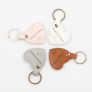 Op de afbeelding: Vier hartvormige sleutelhangers in roze, wit, grijs en bruin. Elke sleutelhanger heeft een metalen ring en een leren band. De roze sleutelhanger zegt "Je t'aime ma soeur". De witte sleutelhanger zegt "Maman d'amour". De grijze sleutelhanger zegt "Meilleur papa". De bruine sleutelhanger zegt "Famille en OR".