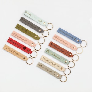 Può includere: Una collezione di portachiavi personalizzati in vari colori, tra cui azzurro, grigio e rosso. Ogni portachiavi presenta un cinturino in pelle con testo inciso e un portachiavi in metallo. Il testo include nomi e frasi come "Romain" e "Chez toi ∞ Chez moi."
