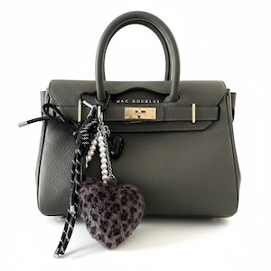 Puede incluir: Un bolso de mano de cuero gris con asa superior y un charm en forma de corazón. El bolso tiene herrajes dorados y la marca "MAC DOUGLAS" es visible en la parte delantera.