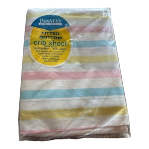 May include: A Penney's fitted bottom crib sheet in 100% cotton fine muslin, packaged in clear plastic. The fabric features horizontal stripes in yellow, pink, and light blue on a white background. The label specifies 166 threads per square inch.