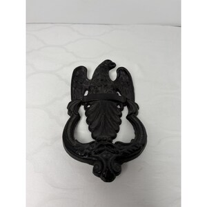 May include: A black cast iron door knocker featuring an eagle with outstretched wings. The eagle is centered above a decorative, symmetrical design. The door knocker is a dark, matte black color and is likely made of metal.