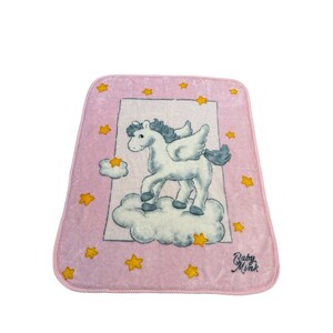 May include: Pink baby blanket featuring a white winged horse standing on a cloud, surrounded by yellow stars. The blanket has a pink border and the words "Baby Mink" are printed on the bottom right corner.