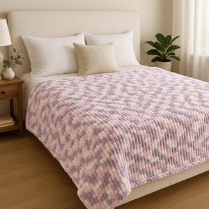 Op de afbeelding: Een zachte, dikke gebreide deken in tinten lavendel, wit en roze ligt op een bed. De deken heeft een getextureerde, pluche uitstraling. Twee witte kussens en een beige kussen zijn zichtbaar.