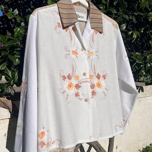 Può includere: Camicia bianca a maniche lunghe con ricami floreali arancioni e marroni. Il colletto presenta strisce marroni e beige. La camicia ha una chiusura con bottoni e il marchio "Reiv" è visibile sul colletto.