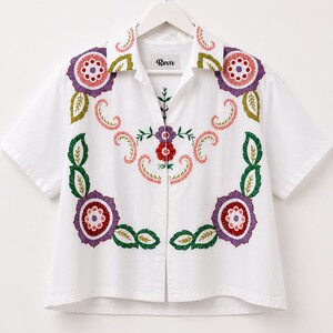 May include: White short-sleeved shirt with colorful floral embroidery. The embroidery features purple, red, pink, and green designs around the collar and front. The shirt has a button-up front and the brand name "Reviv" is visible on the label.