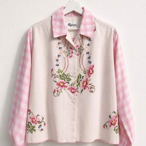 Può includere: Camicia abbottonata color crema con maniche e colletto a quadri rosa. La camicia presenta un motivo floreale a punto croce in rosa, verde e blu. La parola "Reviv" è stampata sul colletto.