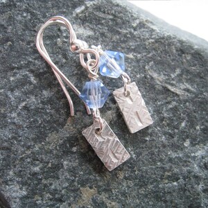 May include: Silver earrings with a textured rectangular charm and a light blue crystal bead.
