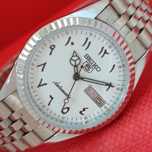 Puede incluir: Reloj de pulsera automático Seiko 5 plateado con esfera blanca y números arábigos. El reloj tiene una pantalla de día y fecha y una correa plateada. La esfera del reloj tiene un bisel texturizado y estriado.