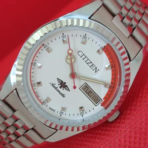 Op de afbeelding: Citizen automatisch polshorloge met een witte wijzerplaat, zilveren wijzers en een rode secondewijzer. Het horloge heeft een zilveren band en een datumweergave. Op de wijzerplaat staan de woorden "Citizen" en "Automatic".