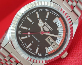 May include: A silver-toned Seiko automatic wristwatch with a dark brown face and a silver band. The watch face features white and orange accents, with the day and date displayed. The watch has a fluted bezel and is set against a red background.