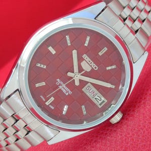May include: A stainless steel Seiko 5 automatic watch with a burgundy dial featuring a textured pattern. The watch has a day and date display, and a silver-toned bracelet. The dial reads "Automatic 17 Jewels" and "Sun 2".