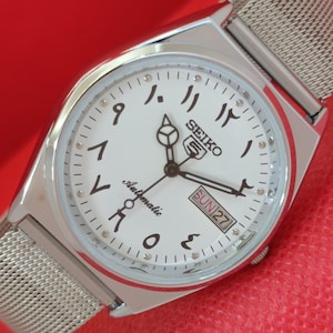 May include: A silver Seiko automatic wristwatch with a white face and Arabic numerals. The watch has a mesh strap and a day-date display showing "SUN 27". The watch hands are black.