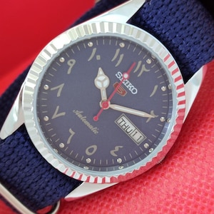 May include: A Seiko automatic wristwatch with a navy blue face and Arabic numerals. The watch has a silver bezel and a navy blue fabric band. The day and date are displayed at the 3 o'clock position. The watch is on a red surface.