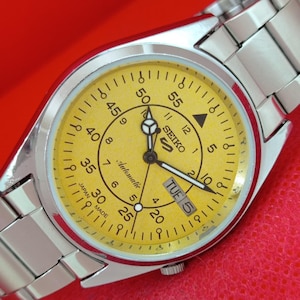 May include: A silver-tone stainless steel wristwatch with a bright yellow dial. The face features black Arabic numerals for both 12-hour and 60-minute markings, the Seiko 5 logo, the word Automatic, and a day-date window displaying TUE 5.