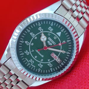 Puede incluir: Un reloj de pulsera automático Seiko de acero inoxidable con esfera verde. El reloj presenta un bisel estriado, un brazalete plateado y una pantalla de día y fecha. La esfera incluye marcadores de hora blancos, agujas luminosas y el texto "SEIKO" y "Automatic".