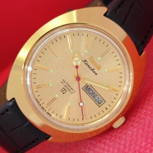 May include: Gold-coloured wristwatch with a light gold face, black leather strap, and a gold bezel. The watch face displays the day and date, with the text "Konchon" and "Automatic 21 Jewels". The watch has a red second hand and green hour markers.