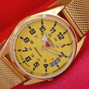 May include: A gold-tone Seiko 5 automatic wristwatch with a yellow dial featuring a geometric pattern, black Arabic numerals, and a 24-hour scale. The watch has a gold-tone mesh strap, luminous hands, and a date window displaying the numbers 28, 29, 30.