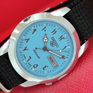 May include: A Seiko 5 automatic wristwatch with a light blue dial featuring Eastern Arabic numerals, silver-tone hands, and a day-date window at the three o'clock position. The watch has a polished silver-tone metal case and a black fabric strap.