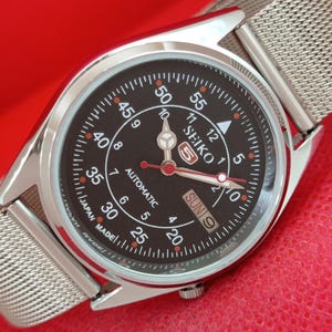 May include: A Seiko 5 automatic wristwatch with a black dial, silver-tone hands, and a red second hand. The dial features white Arabic numerals for minutes and hours, a day-date window at the three o'clock position, and a silver mesh metal strap.