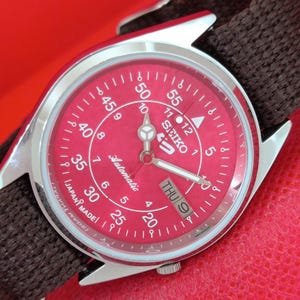 May include: A Seiko 5 automatic wristwatch with a vibrant red dial, featuring white Arabic numerals for both 12-hour and 60-minute markings. The watch has a silver-tone stainless steel case, luminous hands, and a dark brown woven fabric strap.