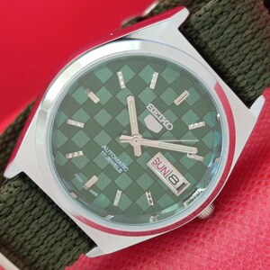May include: A silver-toned Seiko 5 automatic watch with a green checkered dial. The watch features a day and date display, silver hands, and a green fabric strap. The watch face reads "SEIKO 5 AUTOMATIC 17 JEWELS".