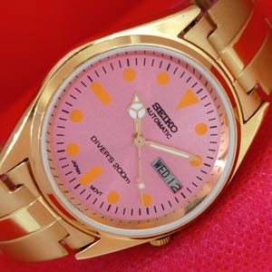 May include: A gold-tone analog wristwatch with a pink dial, featuring orange circular and rectangular hour markers, gold hands, and a day-date window at the three o'clock position. The dial displays the text Seiko Automatic and Diver's 200m.
