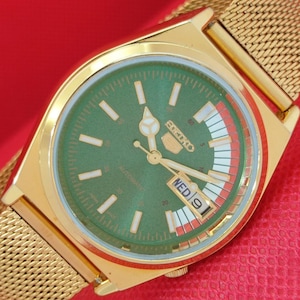 May include: A gold-toned Seiko 5 automatic watch with a green dial and gold hands. The watch features a mesh band and a day-date display. The dial has white hour markers and the text "SEIKO 5 AUTOMATIC".
