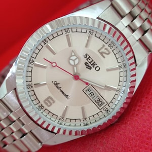 May include: A silver-tone Seiko automatic wristwatch with a round, textured champagne-colored dial, a fluted bezel, and a multi-link metal bracelet. The dial features silver hour markers, a day-date window at the three o'clock position, and a red second hand.