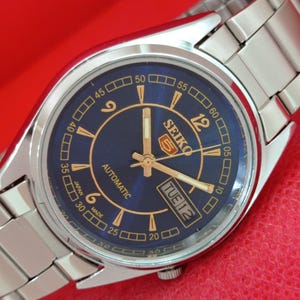 May include: A silver-tone stainless steel Seiko 5 automatic wristwatch with a round blue dial, gold-tone Arabic numerals, and gold-tone hands. The dial features the Seiko 5 logo, the word AUTOMATIC, and a day-date window displaying TUE 12.