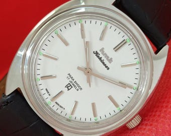 May include: Silver-toned wristwatch with a white face, black leather band, and the text "hmt Kohinoor" in black. The watch face features silver hands and markers, with green accents. The watch is a classic design.