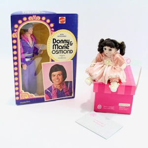 May include: A vintage Donny & Marie Osmond doll in its original box, and a Marie Osmond collectible doll. The Donny doll is in a purple outfit. The Marie doll is in a pink dress, sitting on a pink box.