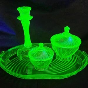 May include: A vibrant green glass vanity set comprising a candlestick, two lidded jars, and an oval tray. The set has a spiral design and is lit against a dark backdrop. The candlestick is approximately 15 cm tall.