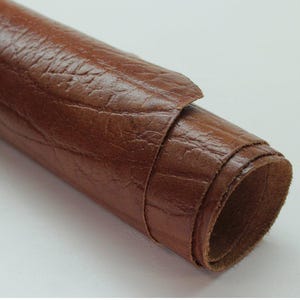 May include: Rolled brown leather material with a textured surface. The leather is a warm, medium brown color and appears to be flexible. The roll is partially unrolled, revealing the interior layers.