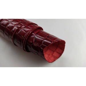 May include: Rolled burgundy leather with a crocodile skin pattern. The leather is a deep red color with a glossy finish. The underside of the leather is a lighter shade of red. The leather is tightly rolled.