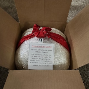 May include: A wrapped ball game inside a cardboard box. The ball is wrapped in clear plastic and tied with a red ribbon. A small white card with black text reads "Treasure Ball Game" and instructions. A die is attached to the ribbon.