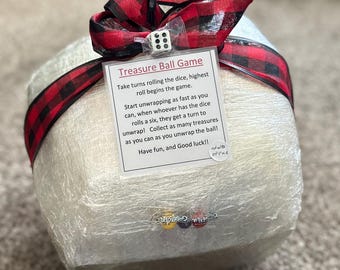 May include: A white, wrapped treasure ball game with a red and black checked ribbon. The game instructions are printed on a white card attached to the ball. The instructions read: "Treasure Ball Game. Take turns rolling the dice, highest roll begins the game. Start unwrapping as fast as you can, when whoever has the dice rolls a six, they get a turn to unwrap! Collect as many treasures as you can as you unwrap the ball! Have fun, and Good luck!!"